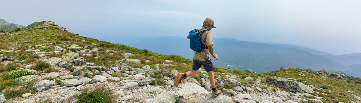 A hiker wearing a beige cap, tan shirt, and dark shorts walks along a rocky mountain trail with a blue backpack, overlooking a vast mountain range under a hazy sky.