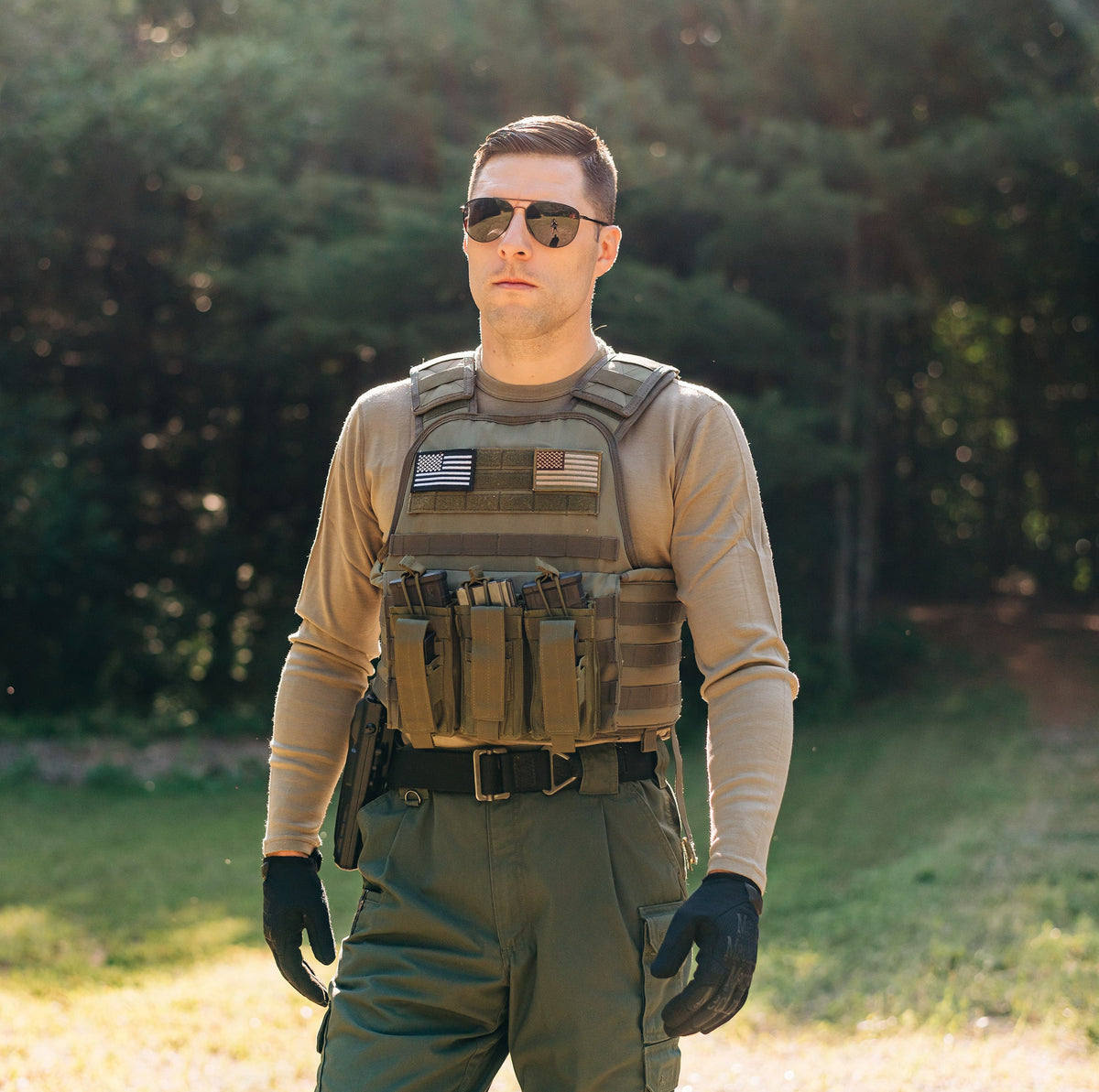 Man standing in a merino wool long sleeve shirt and a bullet proof vest. He has sunglasses on and black gloves and green pants