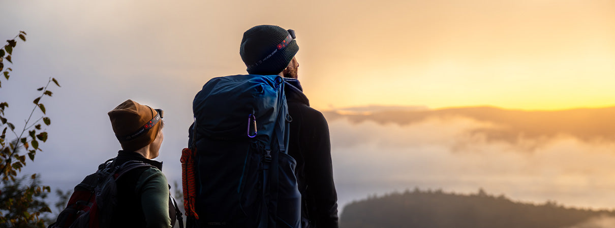 A man and women at the top of a mountain looking up at the sunrise. Their view is foggy and they are wearing merino wool clothing 