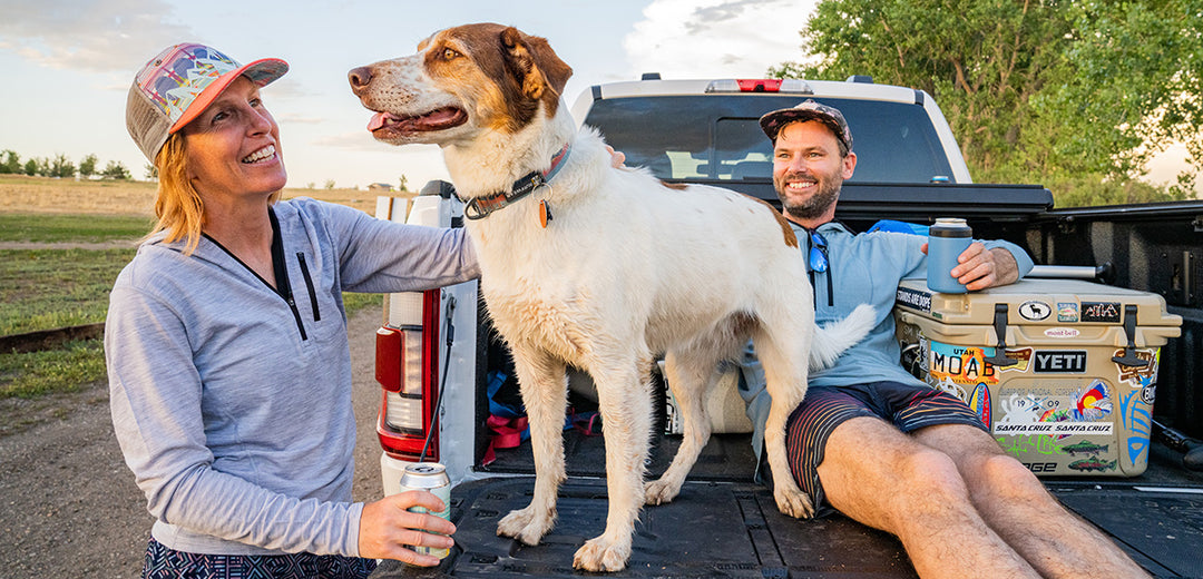 Couple petting their dog on the back of a pickup truck wearing micro weight merino wool sun hoodies in the colors ash gray and slate blue