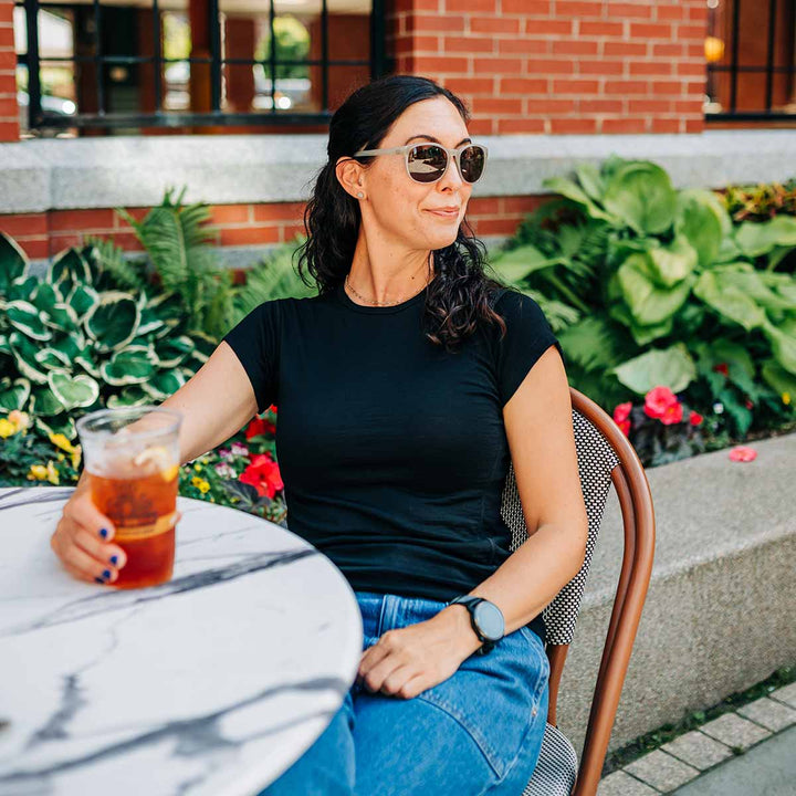 Woman sitting outdoors with a drink, wearing sunglasses and a black merino wool shirt. #color_black
