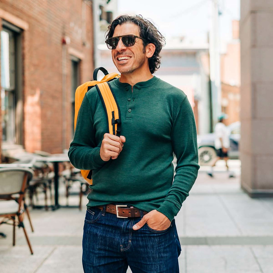 Man wearing a green merino wool sweater and sunglasses, holding a yellow backpack in an urban setting. #color_forest-green