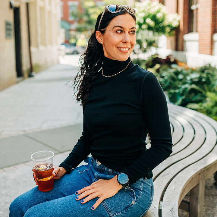 Woman sitting on a bench holding a drink, wearing a black merino wool turtleneck sweater and blue jeans, with a blurred outdoor background. #color_black