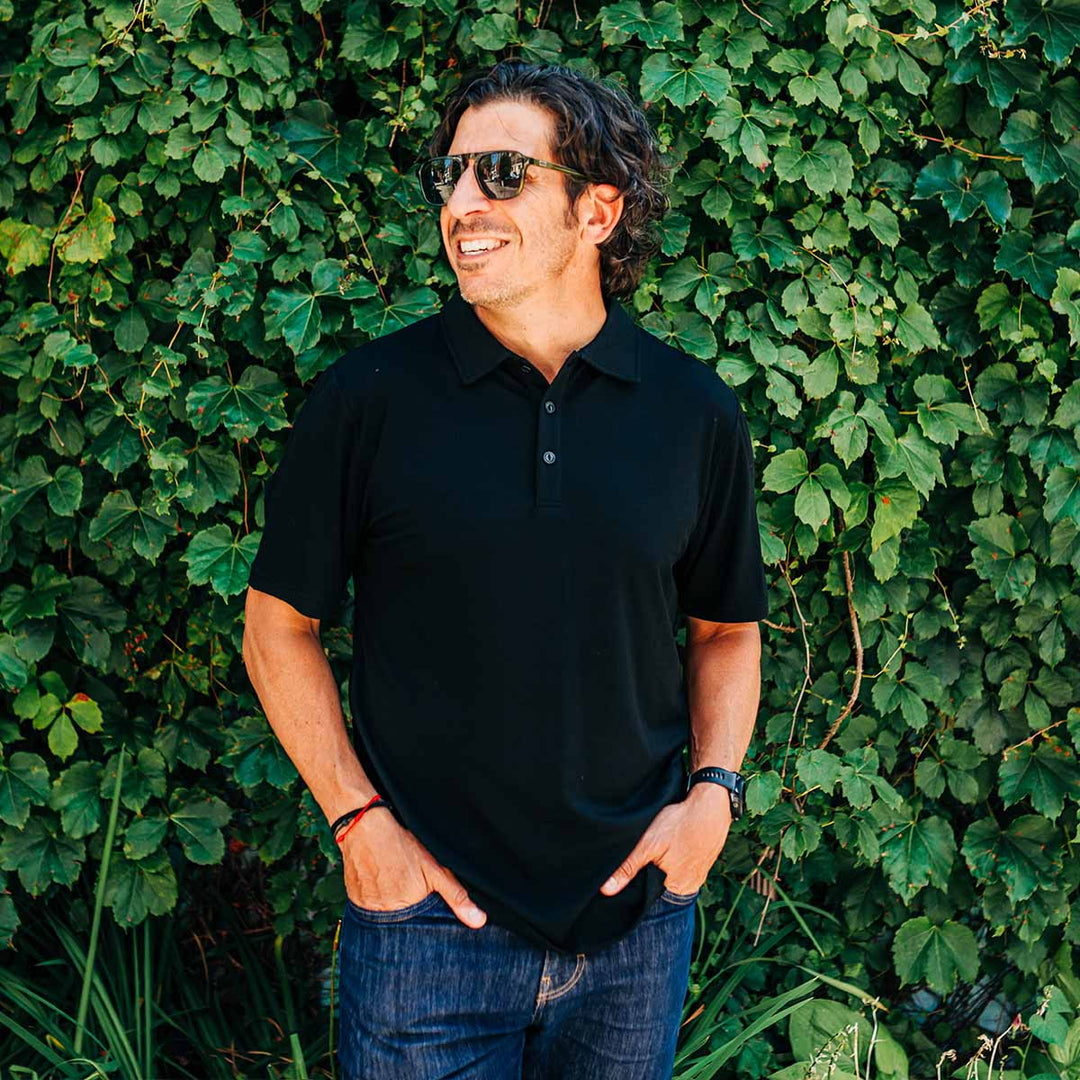 Man wearing a black polo shirt and sunglasses standing against a green leafy background wearing a merino wool polo #color_black