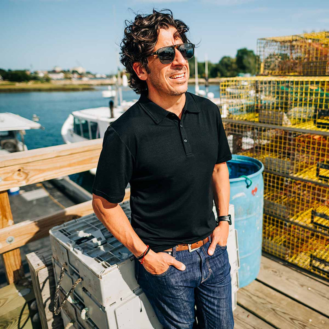 Man in black shirt and sunglasses standing on a dock with lobster traps in the background wearing a merino wool polo #color_black