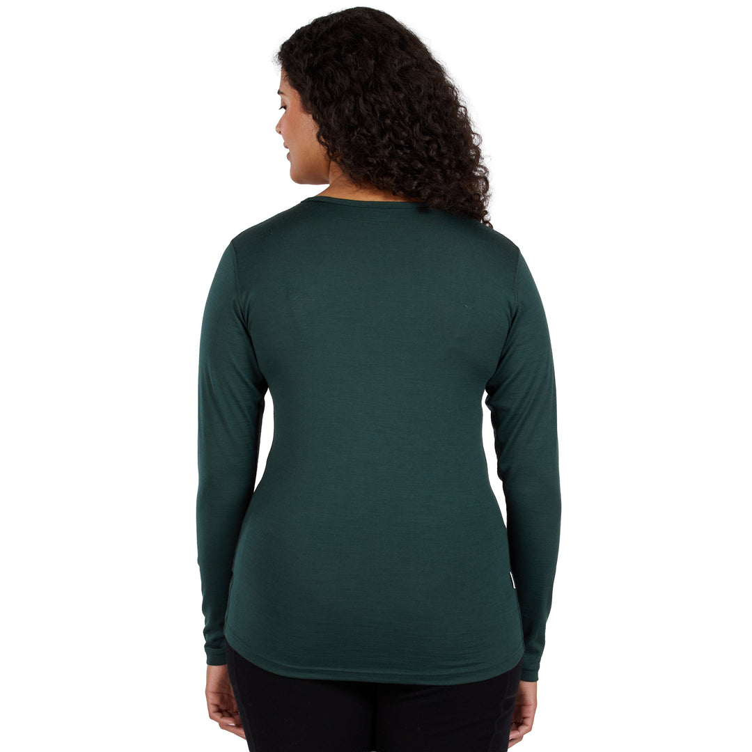 A woman with curly dark hair is shown from the back wearing a dark green 100% merino wool long-sleeve crewneck shirt and black pants against a plain white background. 