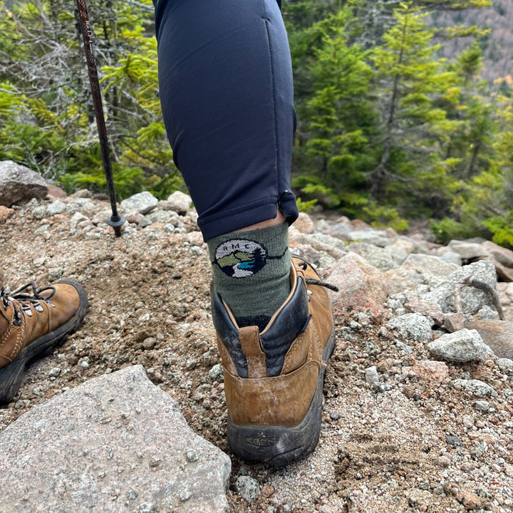 Hiking boot with a sock featuring a mountain logo on a rocky trail. AMC hiking sock