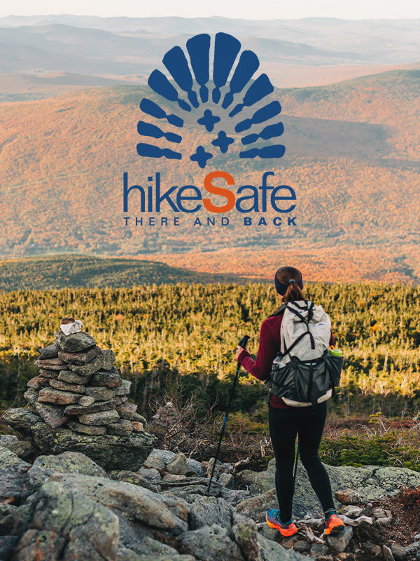 A hiker stands on a rocky mountain trail overlooking a vast autumn forest and hills, with a large 'hikeSafe THERE AND BACK' logo overlaid at the top center.