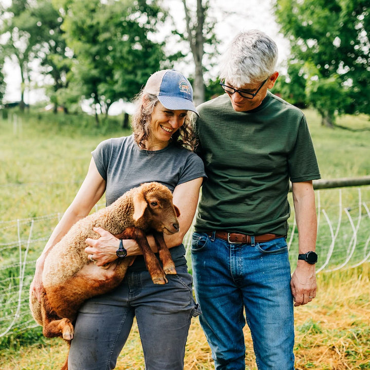 Two people standing in a field with a small lamb, surrounded by trees and greenery. in lightweight merino wool tee shirts #color_olive-drab-green