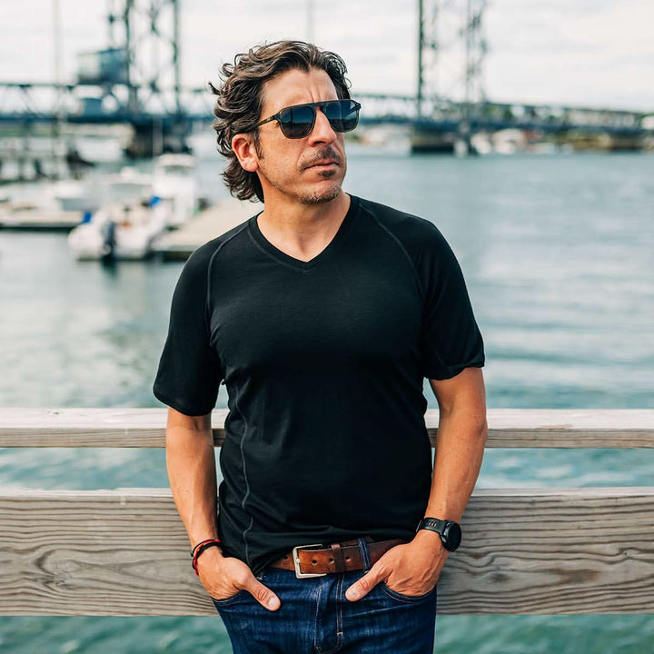 Man wearing sunglasses and a black merino wool t-shirt by a waterfront with boats and a bridge in the background. #color_black