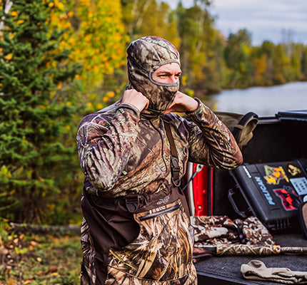 A man wearing a full-body camouflage hunting suit and hood stands in the bed of a truck outdoors near trees and water, with gear visible around him.