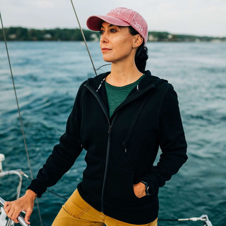 Woman on a sailboat wearing a black wool fleece jacket hoodie and pink cap against a water background. #color_black